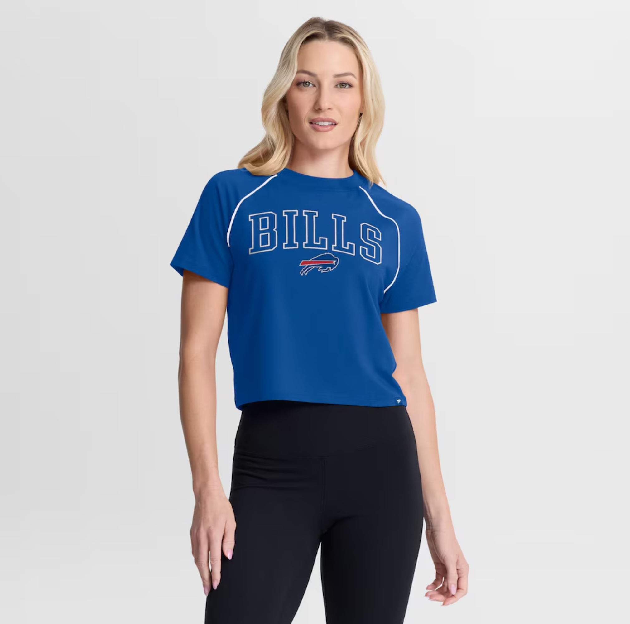 A woman modeling a royal blue short-sleeve athletic crop top with 'BILLS' and the Buffalo Bills logo printed on the front, paired with black leggings, against a light gray studio background.