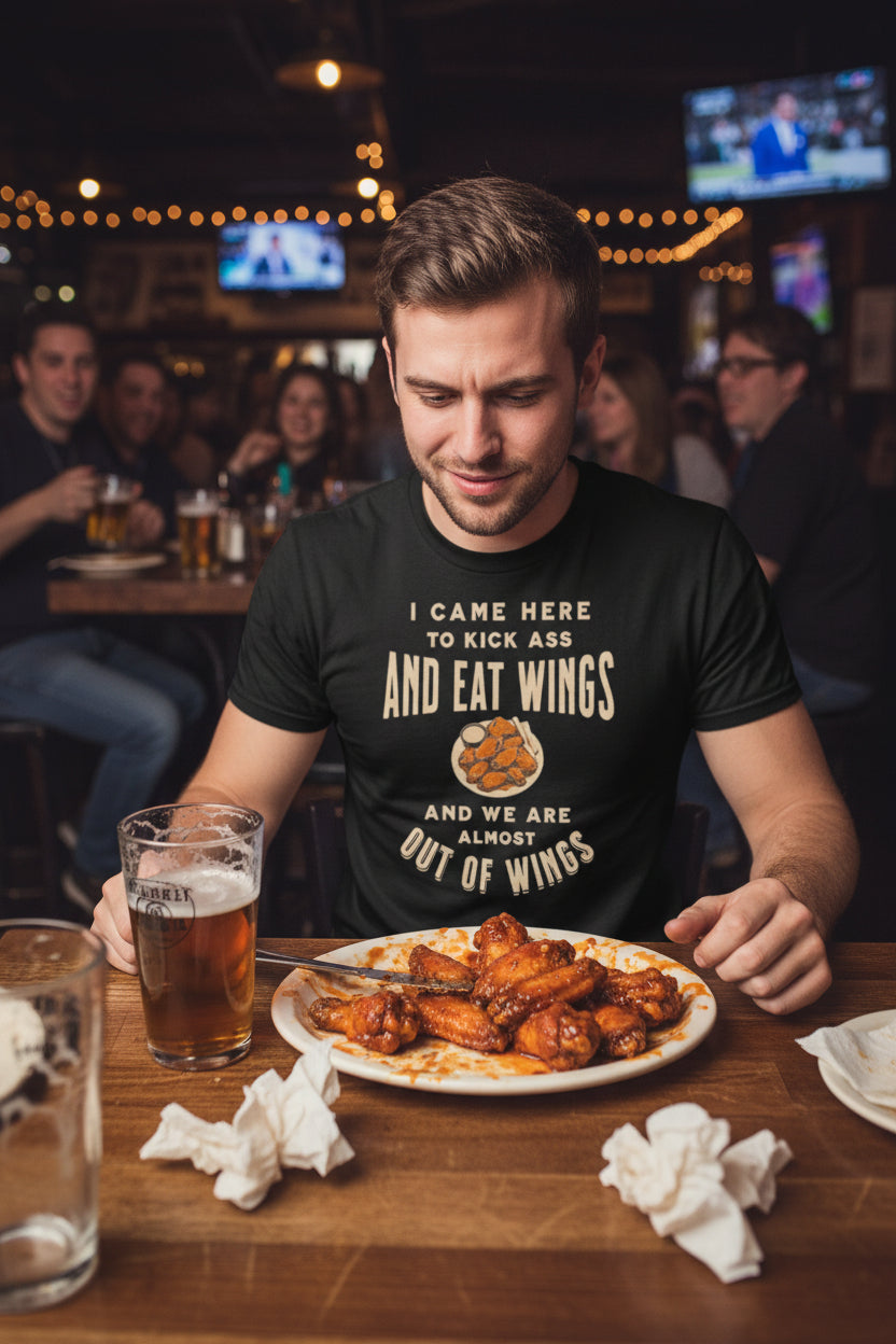 A black short-sleeve crewneck t-shirt with a graphic print on the front. The graphic features the text 'I CAME HERE TO KICK ASS AND EAT WINGS AND WE ARE ALMOST OUT OF WINGS' in a distressed cream font, surrounding an illustration of a winged shoe.